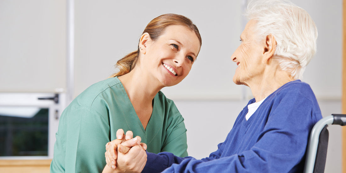 A nurse wearing green scrubs holding hands with an older patient in a wheelchair