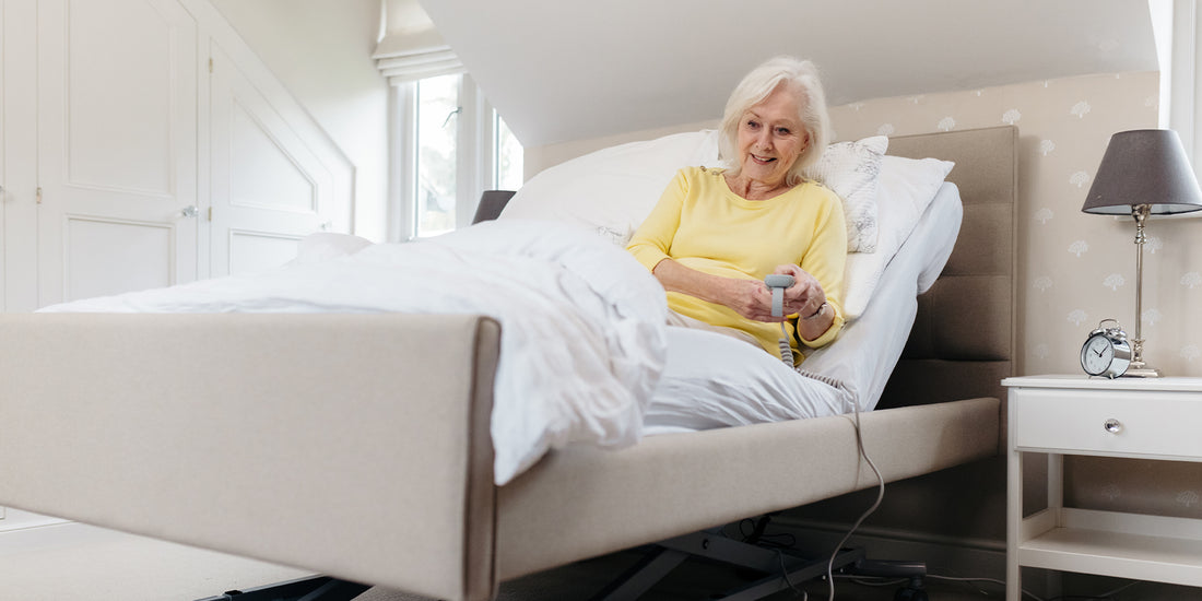 An older woman with blonde hair sat in a profiling bed holding the remote control looking down the bed
