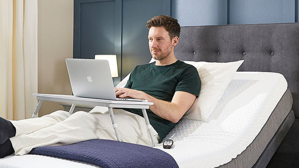 Younger man sitting in an Opera Adjustable Bed using a laptop and lap table.