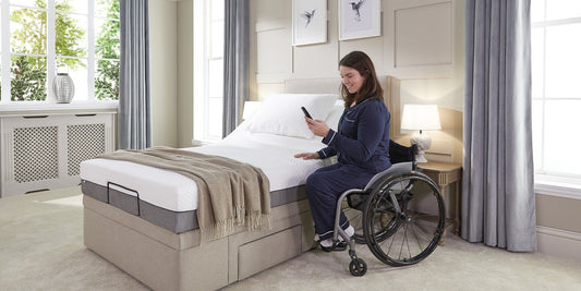 A lady with brown hair sat in her wheelchair next to an Opera adjustable bed holding the wireless control
