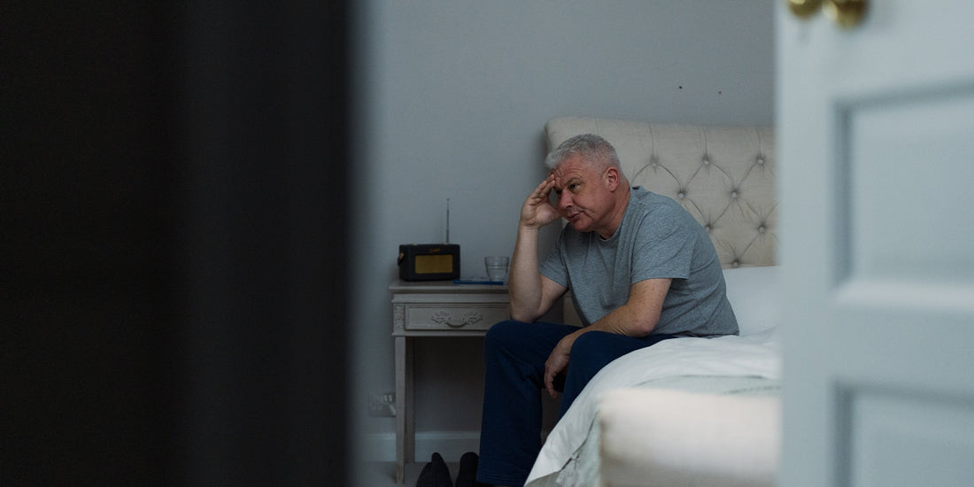 A gentleman sat holding his head in his hands whilst sat at the end of his bed
