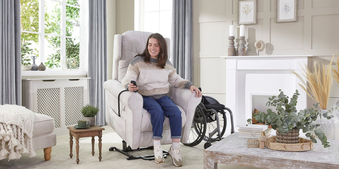 A lady with brown hair sat upright in a Opera riser recliner chair, holding the wired remote with her manual wheelchair in the background