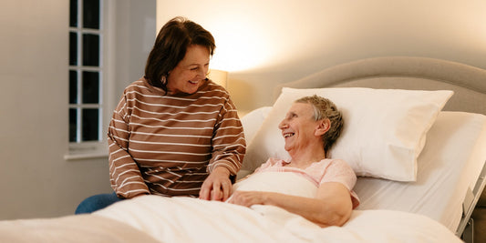 A older lady sat upright in a Opera bed with her daughter sat on the edge of the bed holding her hand