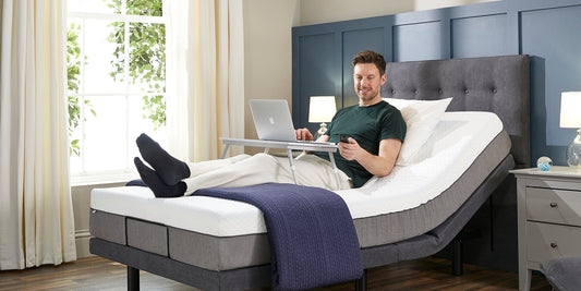 A male sat upright in an adjustable bed holding a wireless control and with a laptop on a table over his legs