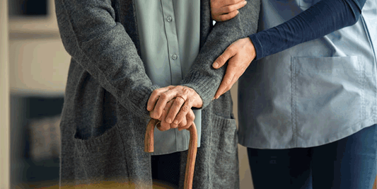 A elderly woman with her hands on a walking stick being supported by a carer