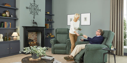 A elderly woman bringing her husband a cup of tea, the gentleman is sat with his feet up in a riser recliner chair