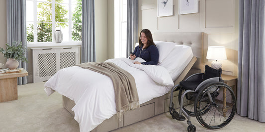 A young female sat upright in an adjustable bed holding the wireless control with her wheelchair next to the bed