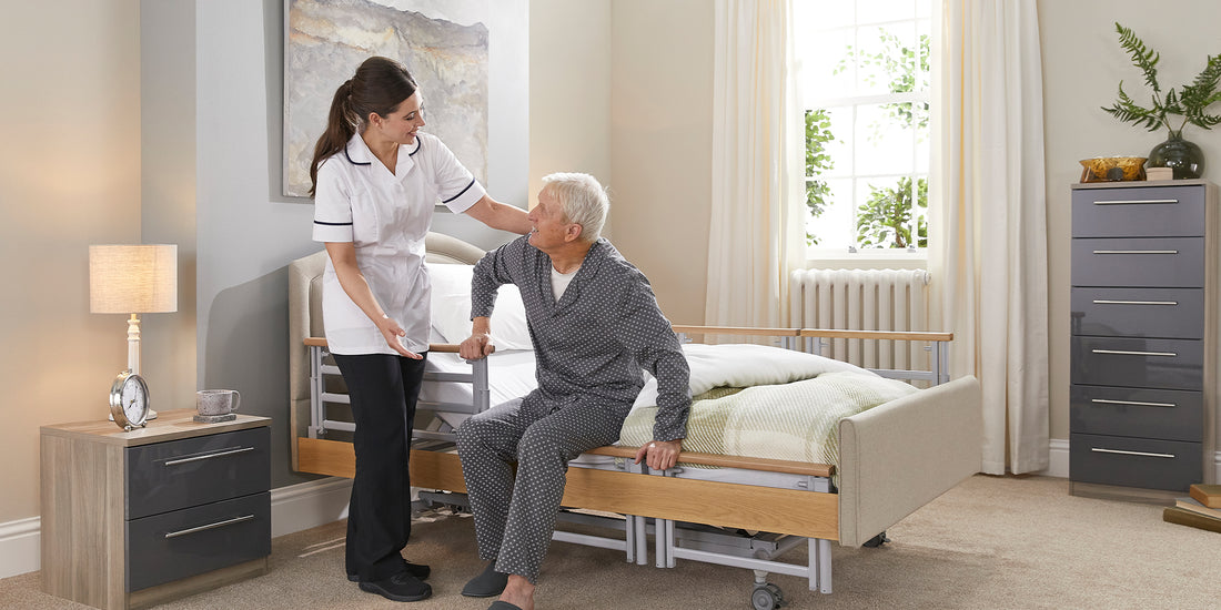 A older gentleman sat on the edge of a profiling care bed with his hands on the side rail and a smiling woman in a carers white uniform holding him and looking at eachother