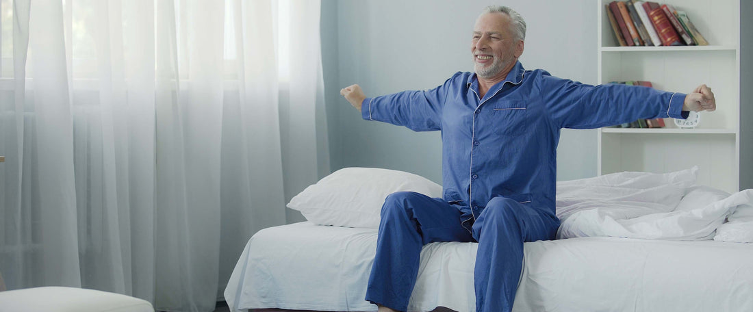 A older gentleman wearing blue pyjamas stretching at the end of his bed