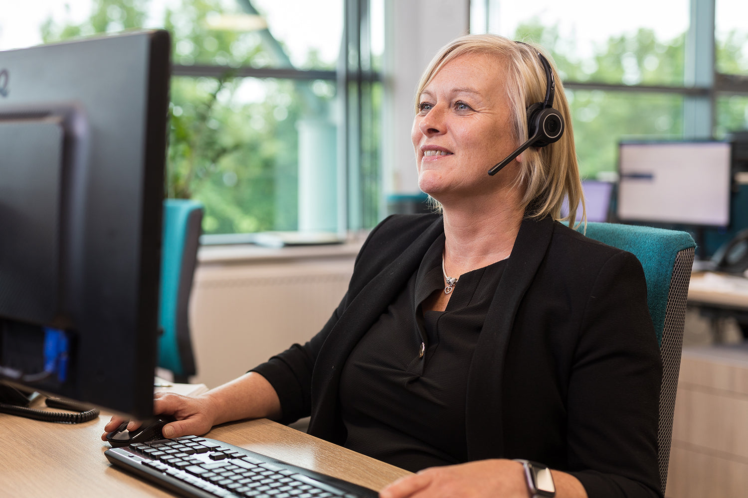 Female customer advisor with blonde hair, wearing a headset and sitting at the computer.