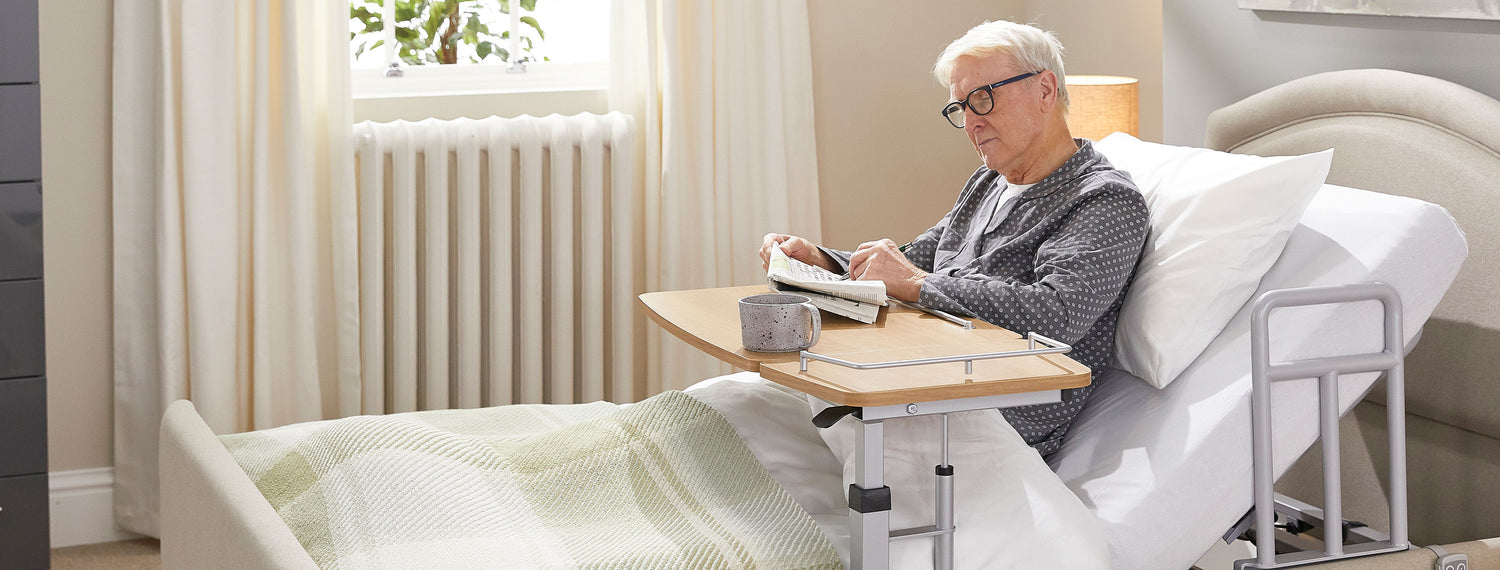Older man wearing glasses and reading a newspaper on an over-bed table. He is sitting in an Opera profiling bed with a grab handle attached to the frame of the bed.