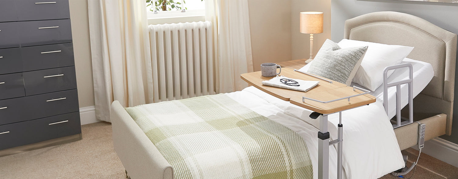 Opera hospital care bed with overbed table with a cup, notebook and pair of glasses on it.