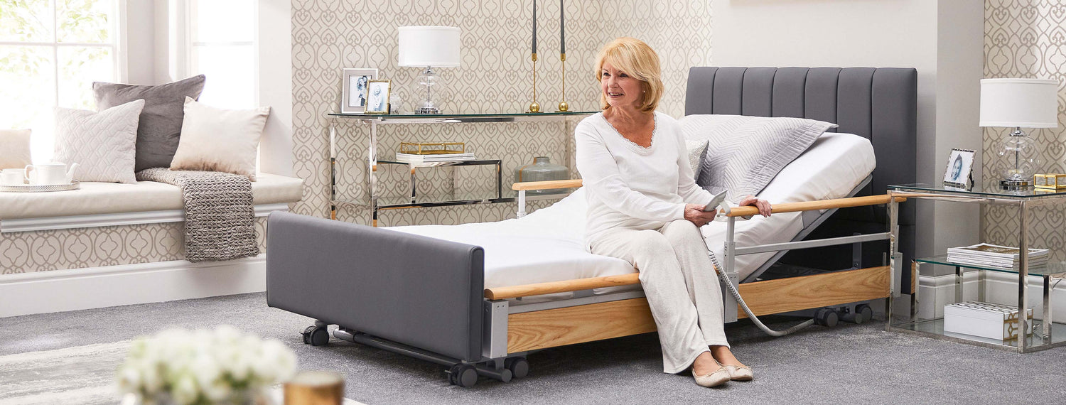 Older woman with blonde hair, sitting on the edge of an Opera profiling bed and using the remote control.