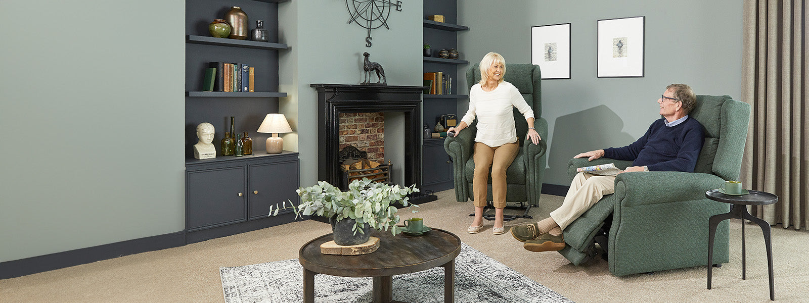 A man and woman both sat in Opera Moss Green Delby riser recliner chairs. The woman is using the tilt then lift function to assist getting out of the chair